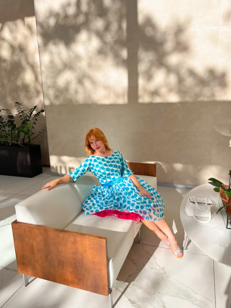 Smiling woman with red hair wearing a blue and white polka dot dress with a pink petticoat, sitting on a modern white couch in a sunlit room.