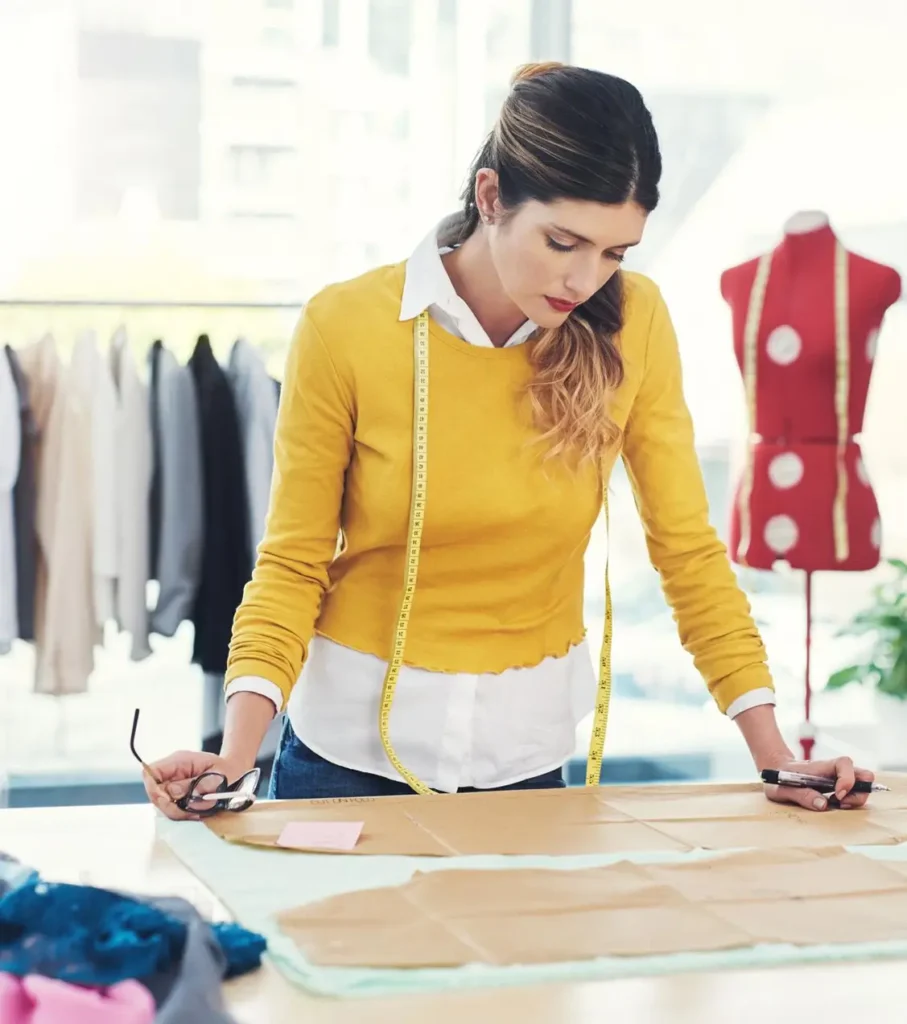 Fashion designer in a mustard-yellow sweater with a measuring tape around her neck leans over a worktable covered in brown paper patterns, holding glasses and a pen, in a bright studio with a red polka-dot dress form and garments on a rack behind her.