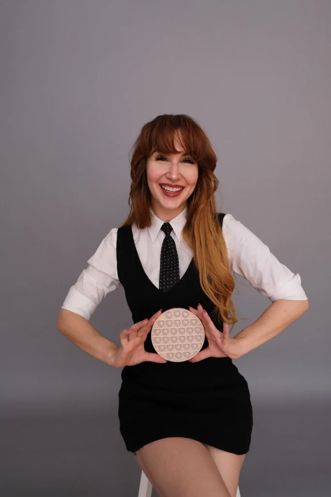 Laughing woman with auburn hair wearing a black dress over a white collared shirt and tie, holding a round pop-it fidget toy against a grey background.