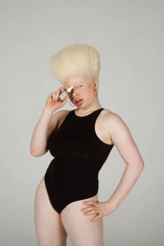 Studio portrait of a Black woman with albinism and a platinum blonde afro, wearing a black bodysuit and holding a skincare dropper bottle to her face.