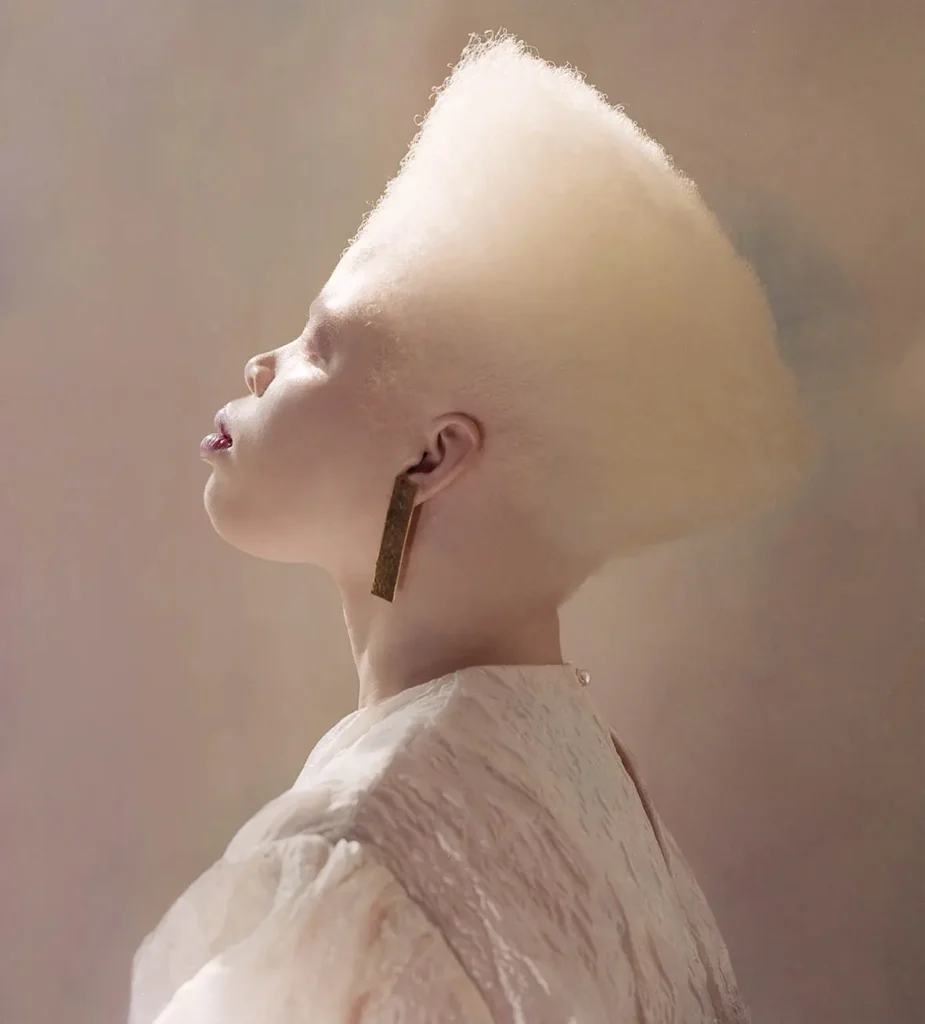 Side profile portrait of a Black model with albinism showcasing a high-top blonde afro, long metallic earrings, and a textured white high-neck blouse.