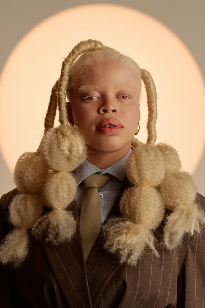 Editorial portrait of a Black person with albinism styling voluminous blonde bubble braids, dressed in a brown pinstripe suit and tie against a glowing circular backdrop.