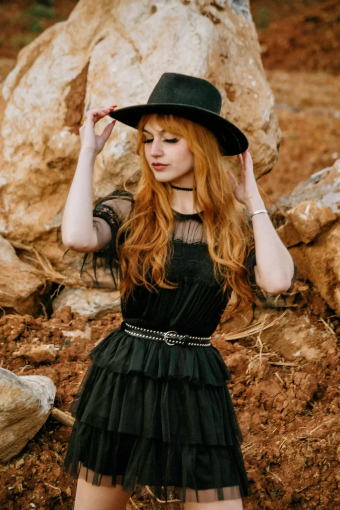 Red-haired woman wearing a black tiered tulle dress and a black wide-brim fedora hat, posing outdoors against large desert rock formations.