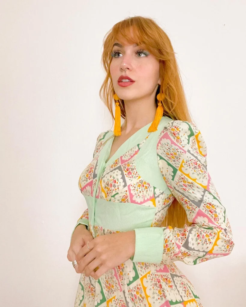 Close-up portrait of a woman with red hair and a nose ring wearing a vintage green pastel floral dress and bright yellow tassel earrings.