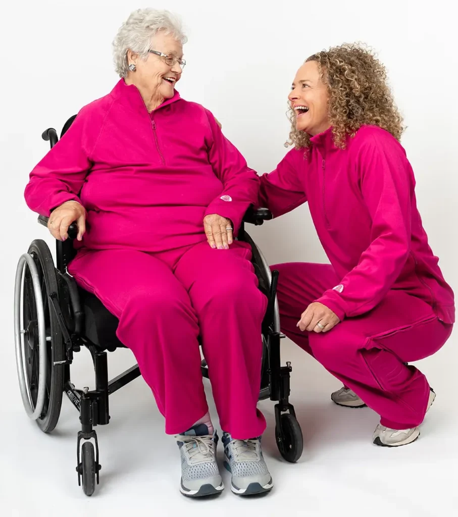 Elderly woman in a wheelchair laughs with a friend, both in pink Adaptt Apparel tracksuits.