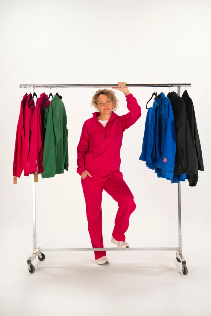 A woman standing in the center of a wide shot wearing a pink Adaptt Apparel tracksuit, leaning against a rolling clothing rack stocked with colorful adaptive hoodies and pants.