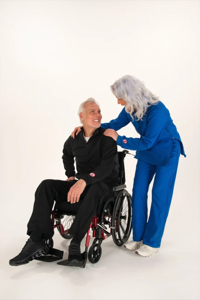Senior man in a wheelchair wearing a black Adaptt Apparel adaptive track suit, smiling at a woman in a blue adaptive outfit against a white background with a left-side gradient overlay.