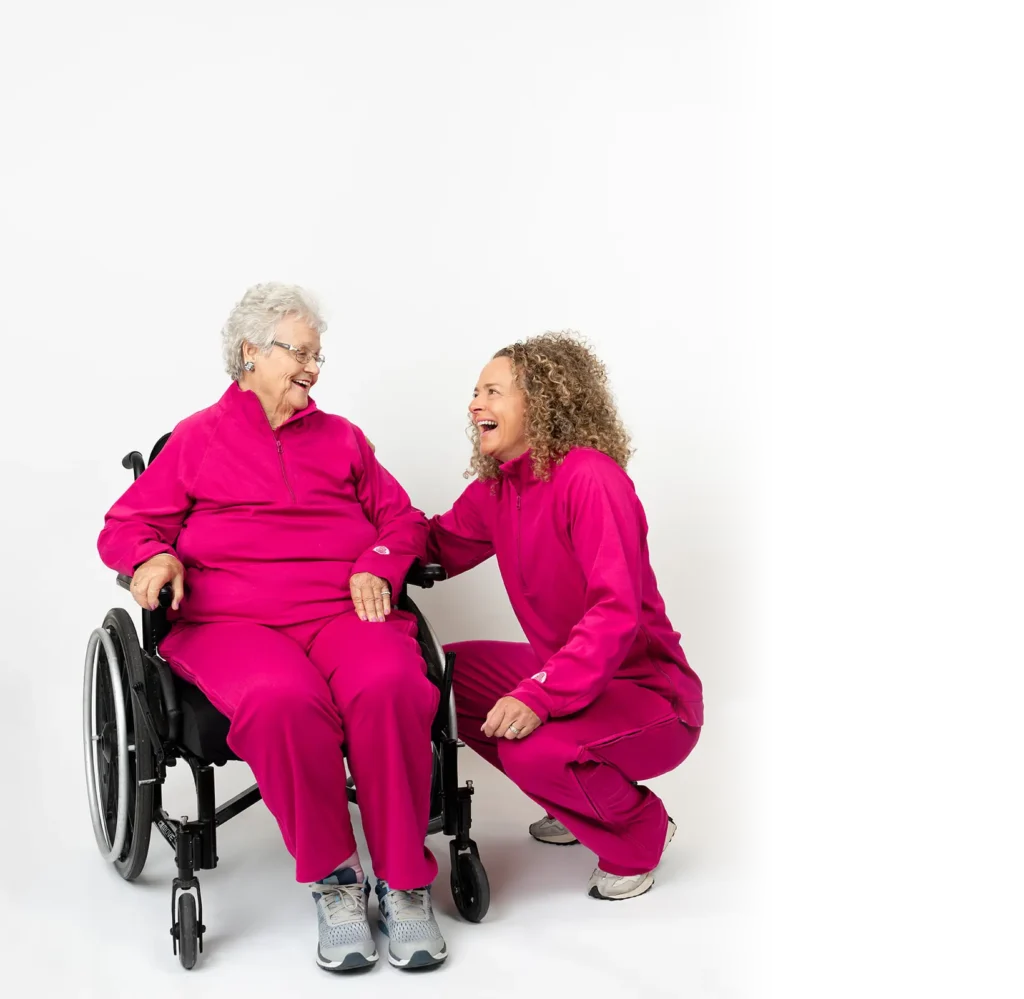 An elderly woman with glasses in a wheelchair laughing with a caregiver or friend, both wearing matching bright pink Adaptt Apparel adaptive fleece tracksuits against a white background.