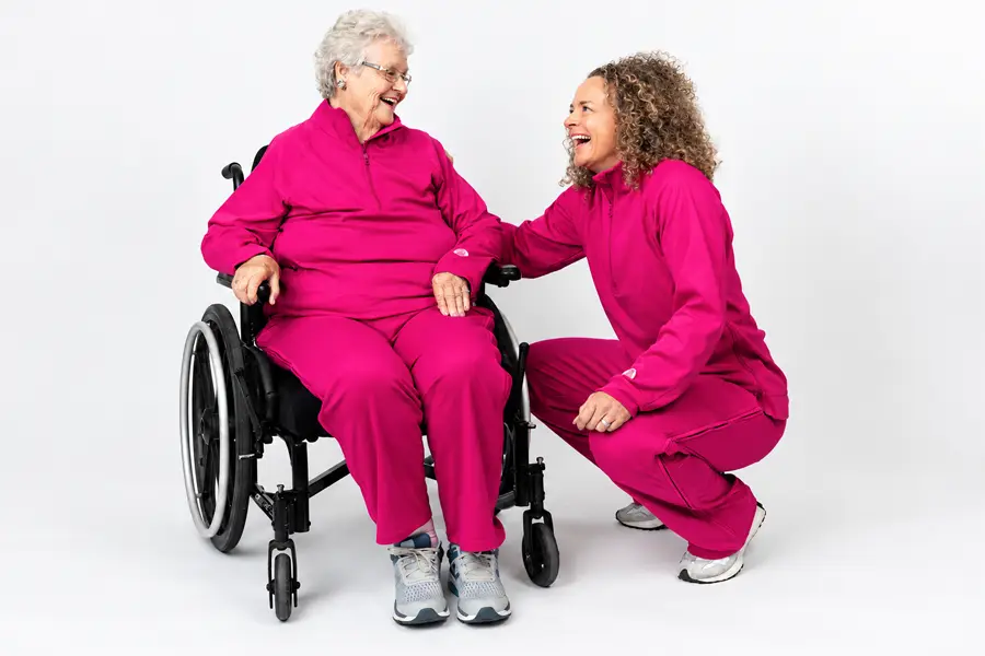 An older woman with short white hair and glasses sits in a black wheelchair, smiling broadly as she looks at the woman beside her. A younger woman with curly, shoulder-length hair crouches next to the wheelchair, laughing, with one hand resting on the older woman’s arm. Both wear matching magenta zip-neck jackets and pants with a small logo on the chest and sleeve, paired with gray and white sneakers. The studio photo has a plain white background.