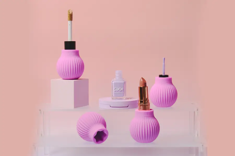 A pink-themed beauty product display featuring ribbed lip gloss bottles, a nail polish bottle, and a lipstick arranged on tiered acrylic risers against a peach background.