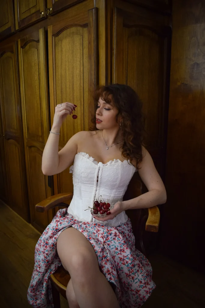 Woman with dark curly hair sitting in a wooden chair, wearing a white lace corset top and floral skirt while holding a small bowl of fresh red cherries.