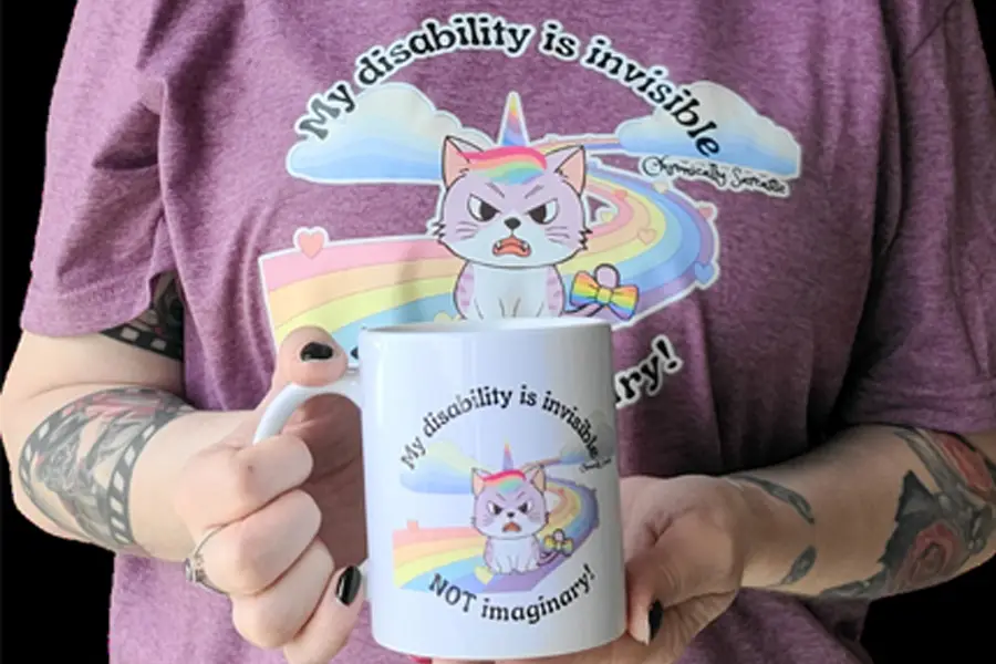A close-up of a person, cropped from the neck down, holding a white ceramic mug with both hands. Both the mug and their heathered purple t-shirt show the same illustration: an angry-looking lavender cat with a rainbow mane and a unicorn horn sitting on a pastel rainbow road, with clouds and a small rainbow bow nearby. Curved text above the design reads ‘My disability is invisible,’ and text below on the mug reads ‘NOT imaginary!’ A small ‘Chronically Sarcastic’ label appears beside the shirt design. The person has black nail polish, a silver ring, and detailed tattoos on both forearms, including a film-strip pattern on the left arm.