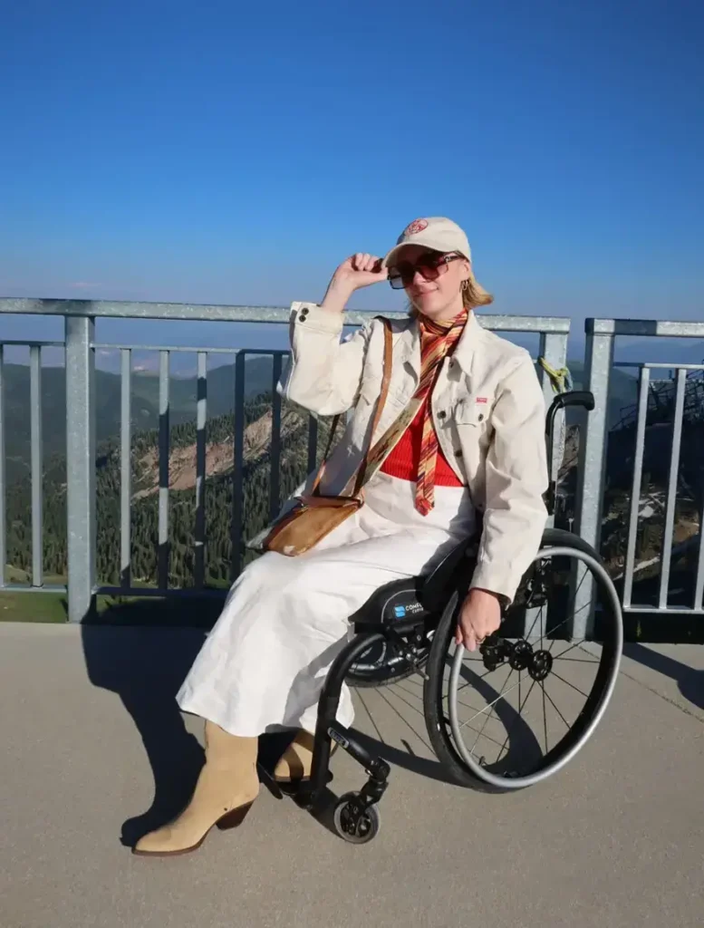 Person in a black wheelchair on a mountain overlook, wearing a cream cap, sunglasses, off-white jacket, red top, striped scarf, long white skirt, and tan boots, with forested mountains and a clear blue sky behind them.