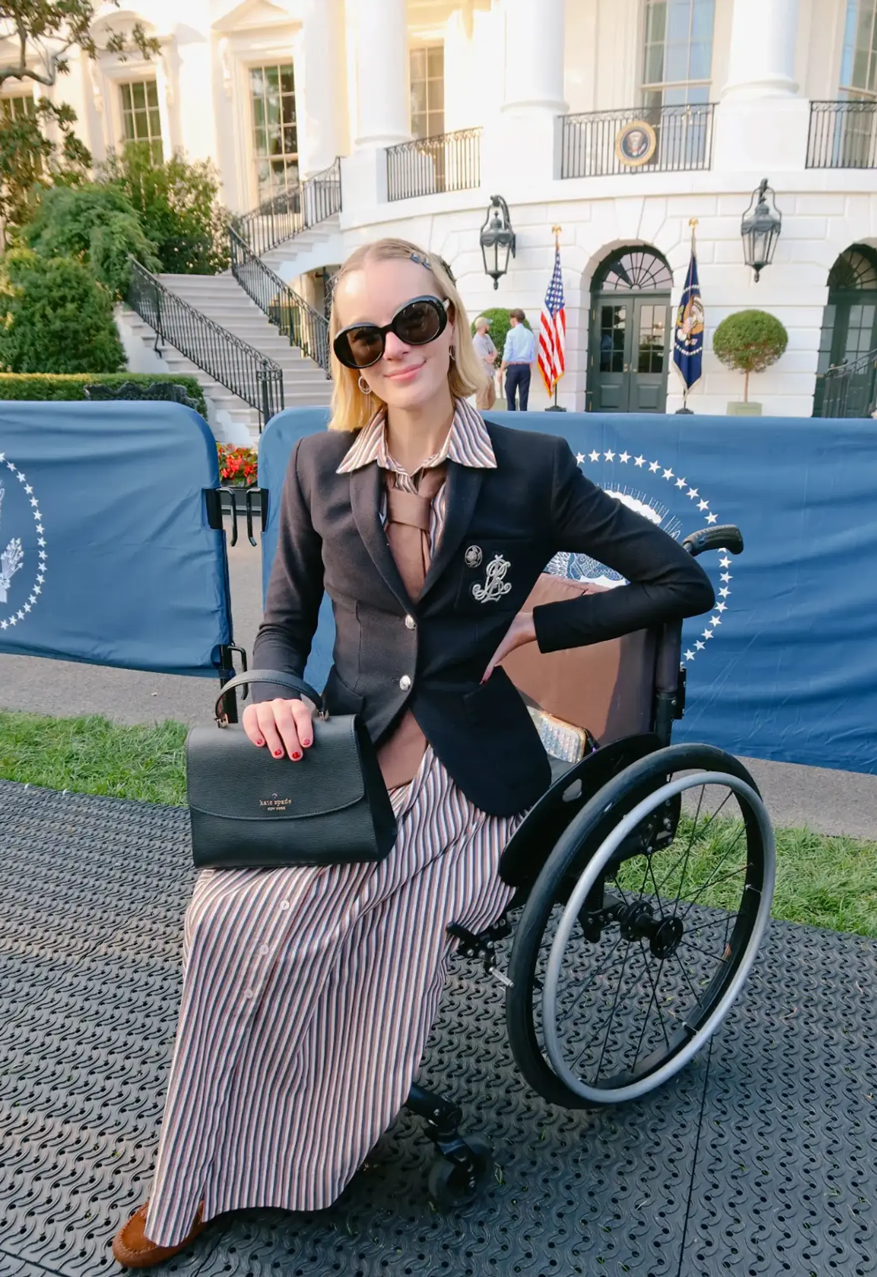 Person with a chin-length blonde bob and oversized dark sunglasses sits in a black wheelchair, wearing a navy blazer over a striped shirt-dress and holding a black handbag, in front of the White House's curved white facade and blue banners with the presidential seal.
