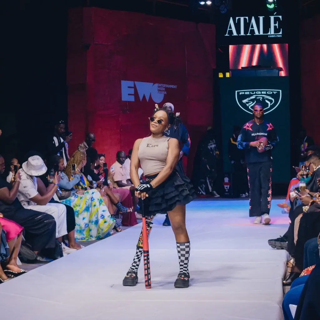 A stylish Black woman striking a playful pose on an indoor fashion show runway for Entertainment Week Ghana (EWG). She is wearing sunglasses, a beige crop top, a tiered black ruffly mini skirt, checkerboard socks, and platform shoes, while leaning on a red baseball bat and showcasing her custom patterned prosthetic leg.