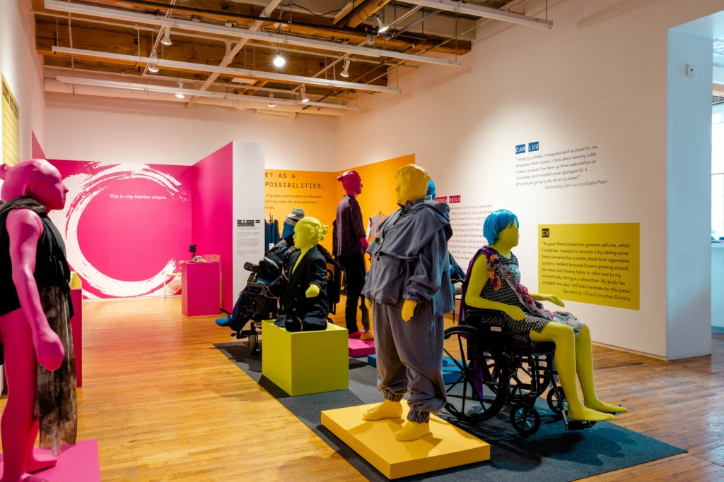 An art gallery exhibition space featuring brightly colored mannequins in pink, yellow, and blue displaying adaptive clothing. One mannequin sits in a manual wheelchair, while text on the white wall reads "This is crip fashion utopia."