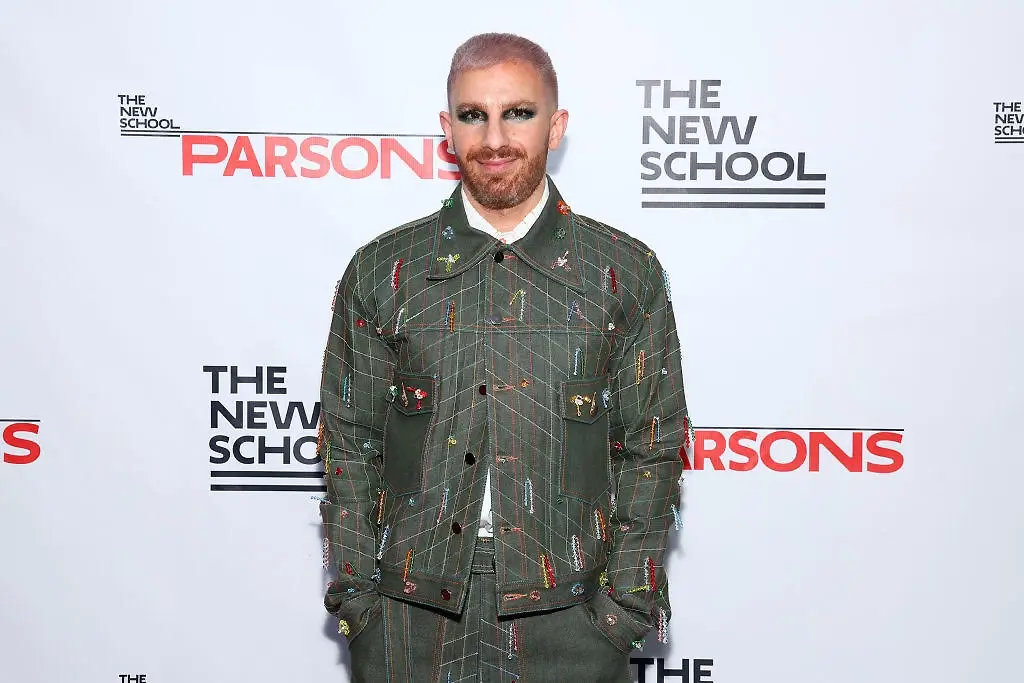 Dr. Ben Barry smiling on a red carpet in front of a white backdrop with "The New School Parsons" logos, wearing a olive green grid-patterned denim jacket adorned with colorful beaded safety pins, paired with blue eye makeup.