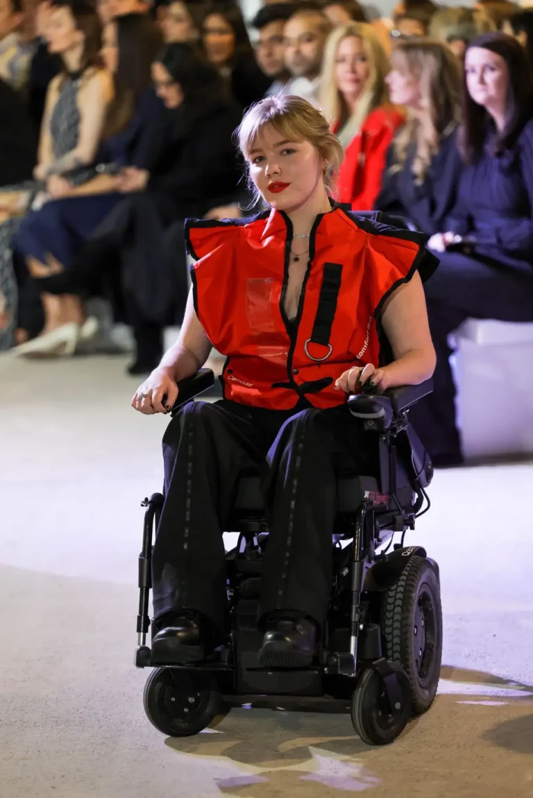 A blonde model with red lipstick modeling a structured red vest and black trousers while driving a black power wheelchair down a fashion show runway with an audience in the background.