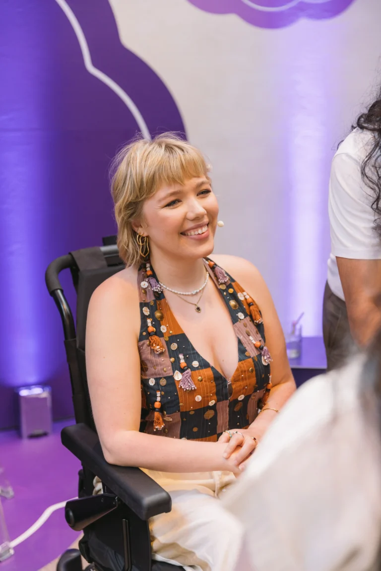 A smiling blonde woman with short hair sitting in a power wheelchair, wearing a patterned halter top, during an indoor panel discussion with a purple background.