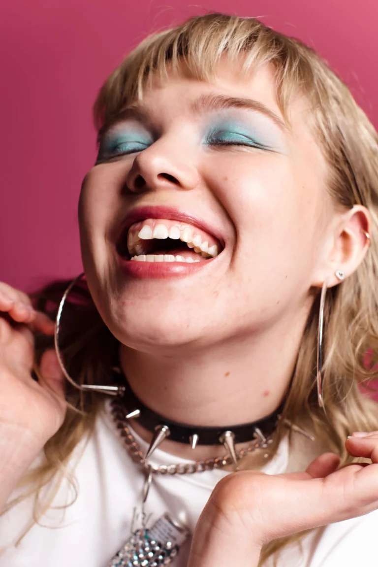 Close-up portrait of a laughing blonde woman with blue eyeshadow, large hoop earrings, and a black spiked leather choker against a vibrant pink background.
