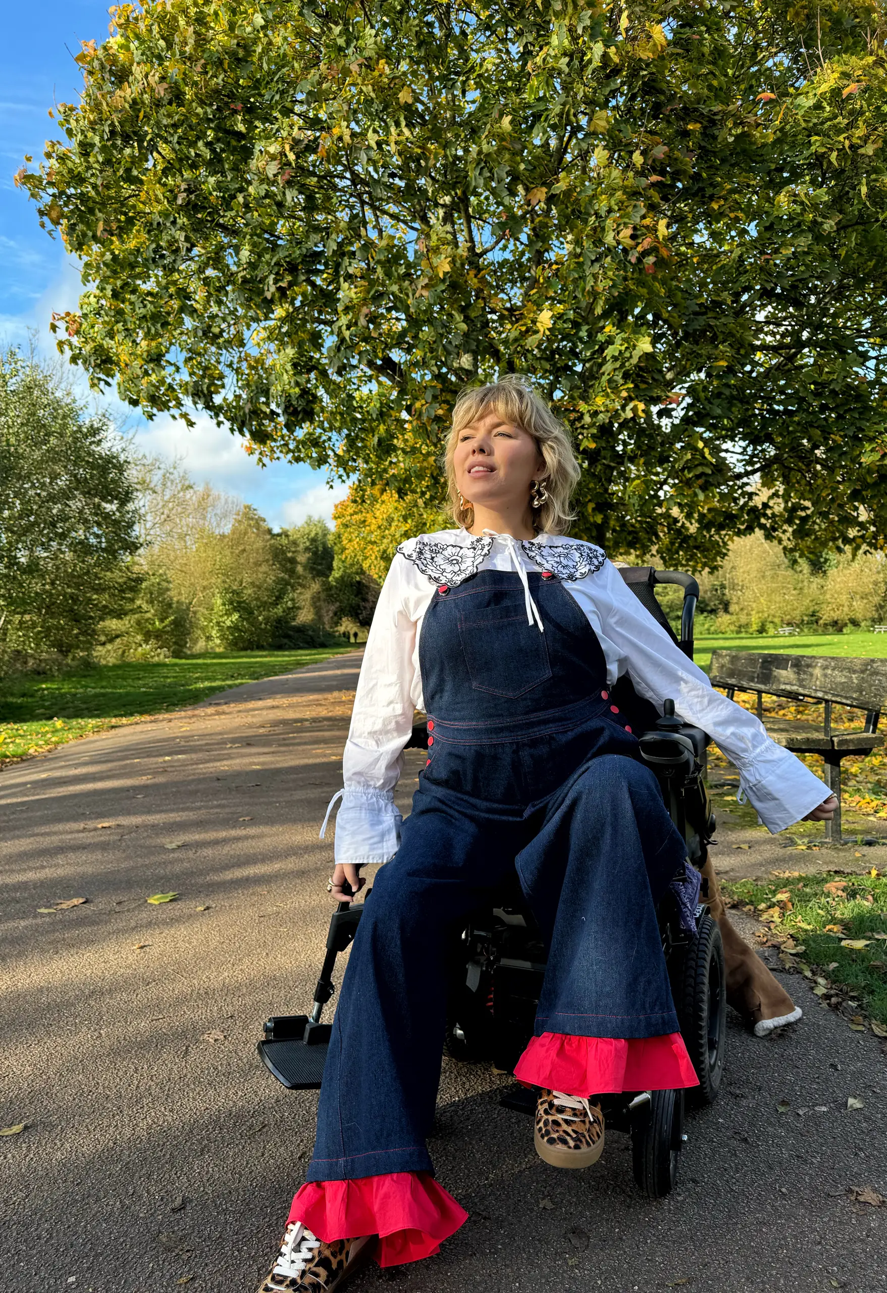 Person with short wavy blonde hair sits in a black power wheelchair on a park path, gazing upward, wearing dark denim overalls with bright pink ruffled hems, a white blouse with an embroidered collar, and leopard-print sneakers, beneath a tree in autumn leaves.