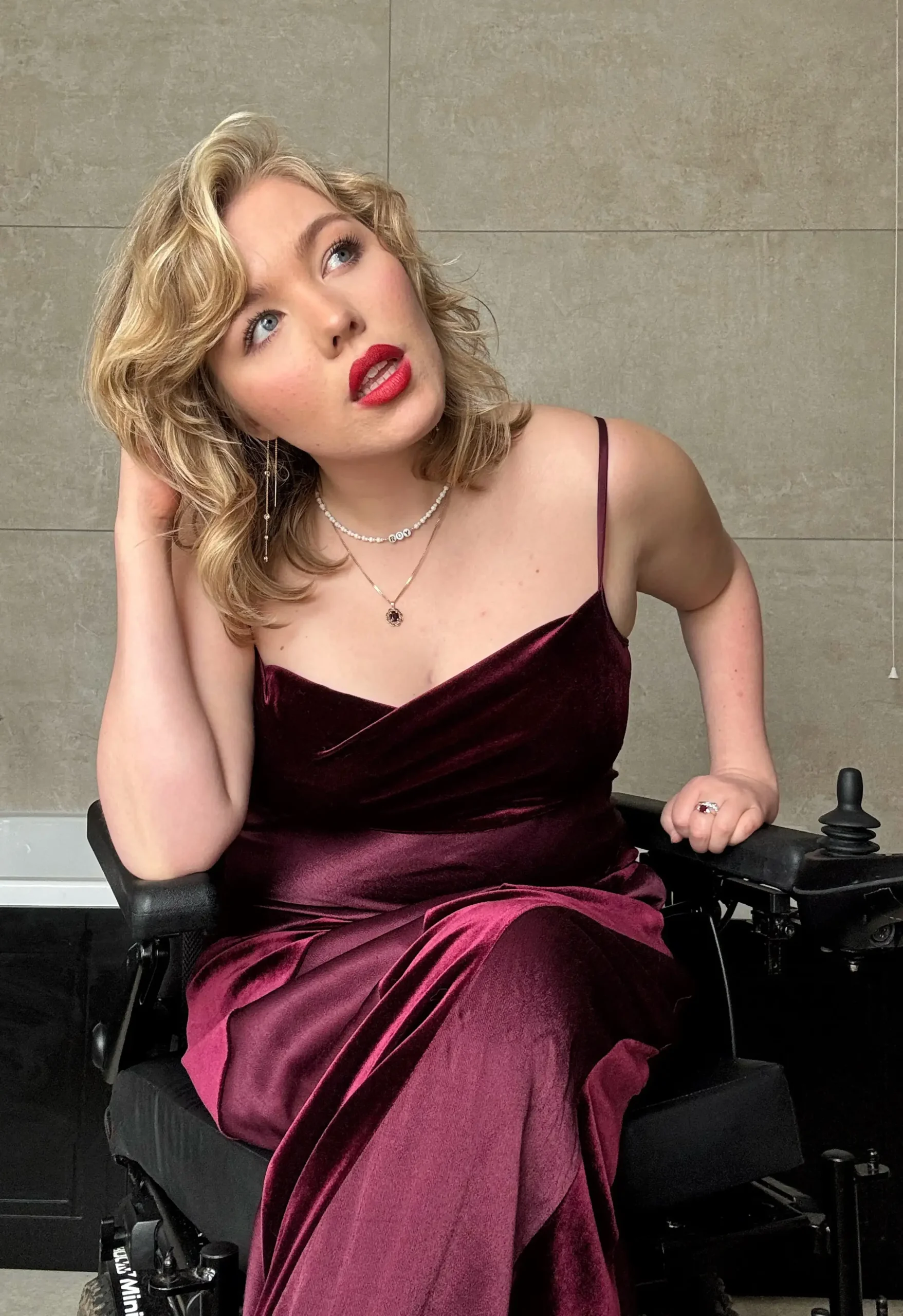 Close-up portrait of a blonde woman with red lipstick wearing a formal burgundy velvet gown while sitting in a motorized wheelchair.