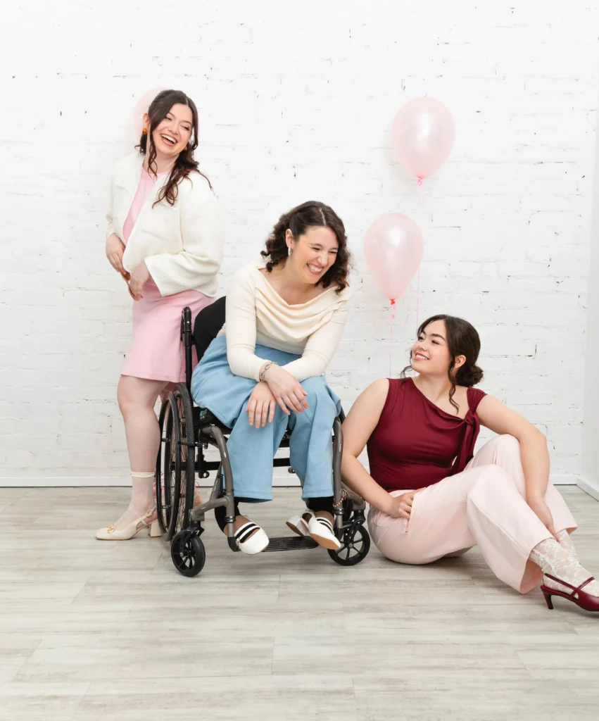 Three cheerful women laughing together in a studio setting with a white brick wall and pink balloons. One woman stands in a pink dress and white jacket; the second sits in a wheelchair wearing a cream top and blue trousers; the third sits on the floor in a maroon top and pink pants.
