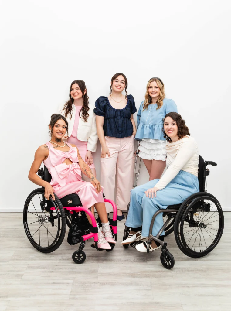 Group portrait of five smiling women showcasing inclusive fashion against a white background, including two women using wheelchairs, one using crutches, and two standing.