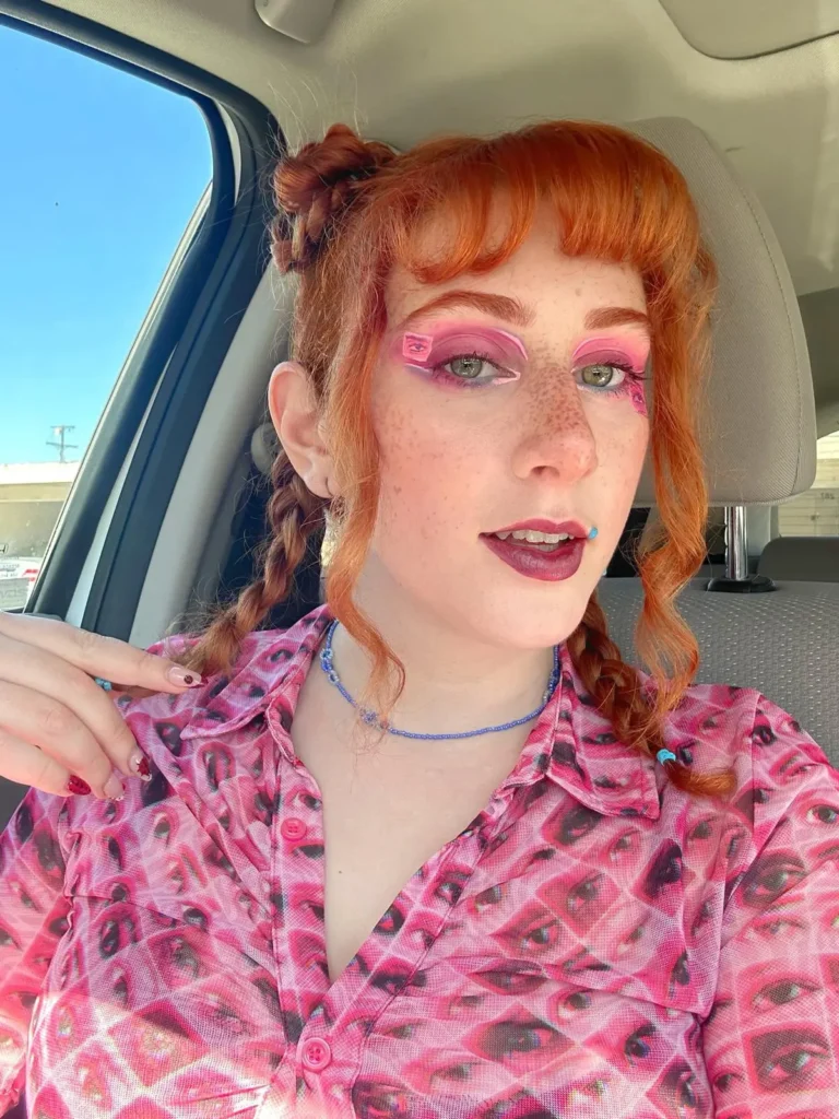 Close-up selfie of a person with bright orange hair, graphic pink eye makeup, and a pink mesh shirt featuring an all-over eye print pattern.