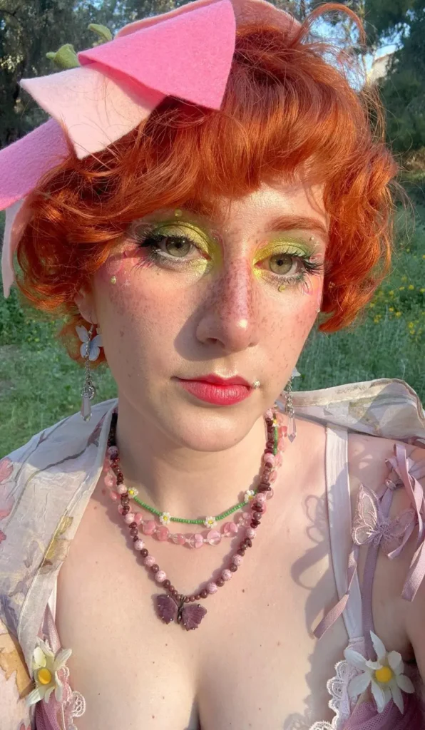 Close-up portrait of a person with curly red hair, fairy-inspired green eye makeup, butterfly necklaces, and a pink felt bow headpiece.