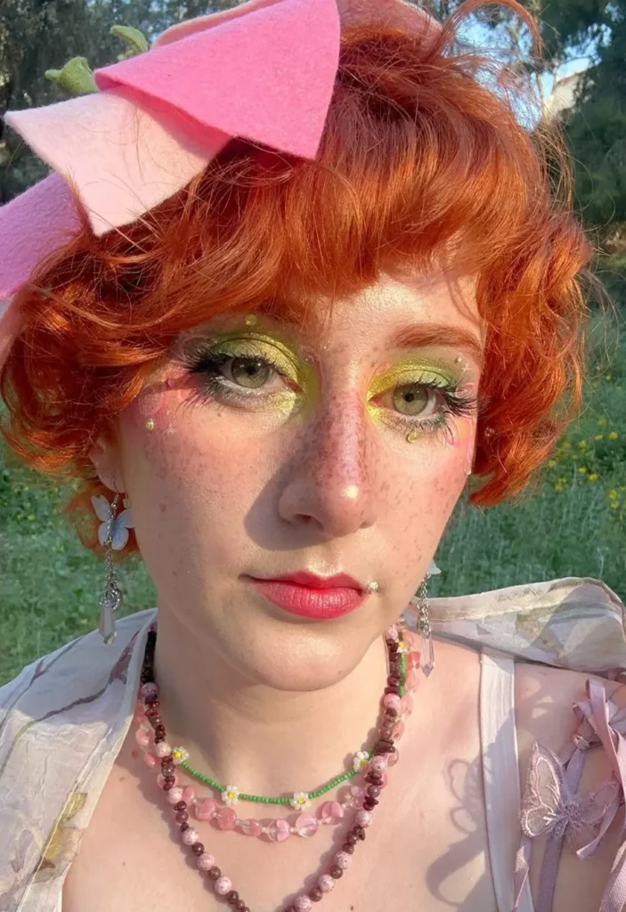 Close-up of a person with curly red hair, green eye makeup, and a pink felt bow headpiece.