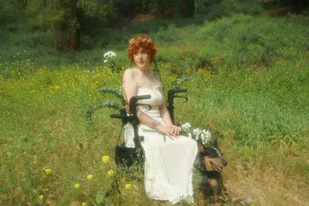 A person with curly, bright red hair wearing a white flower crown and a long white dress sits in a black wheelchair in a grassy field filled with small yellow wildflowers. Eucalyptus branches and white flowers are woven into the handles of the wheelchair, and the person holds a bouquet of white flowers in their lap while looking off to the side. A medium-sized, black-and-tan dog sits loyally on the grass beside the wheelchair, looking towards the right. The background features a lush, green hillside with soft, hazy sunlight filtering through the trees.