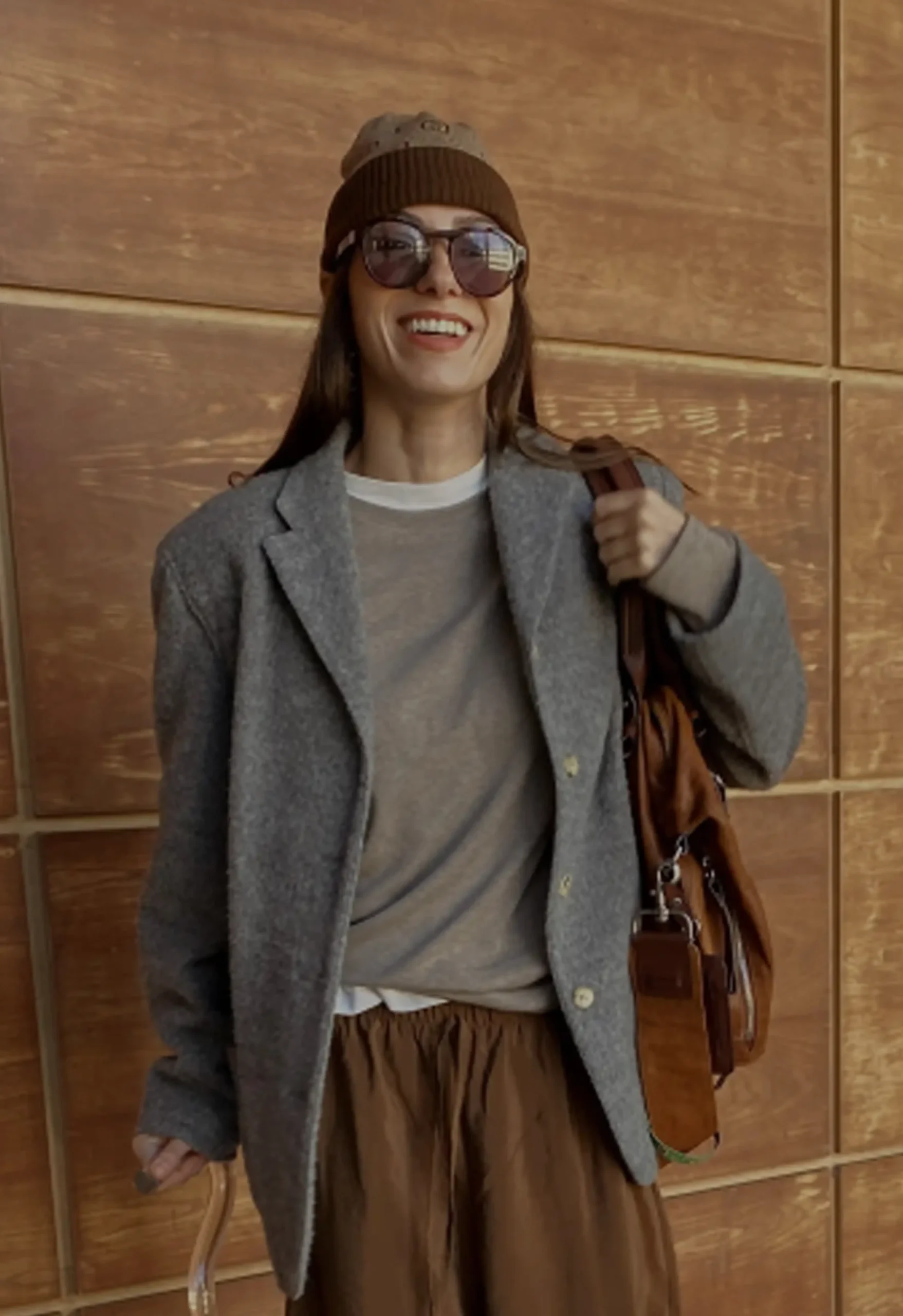 A smiling woman wearing a gray wool blazer, beige t-shirt, brown wide-leg trousers, a knit beanie, and sunglasses walking with a clear acrylic cane while carrying a brown leather bag.