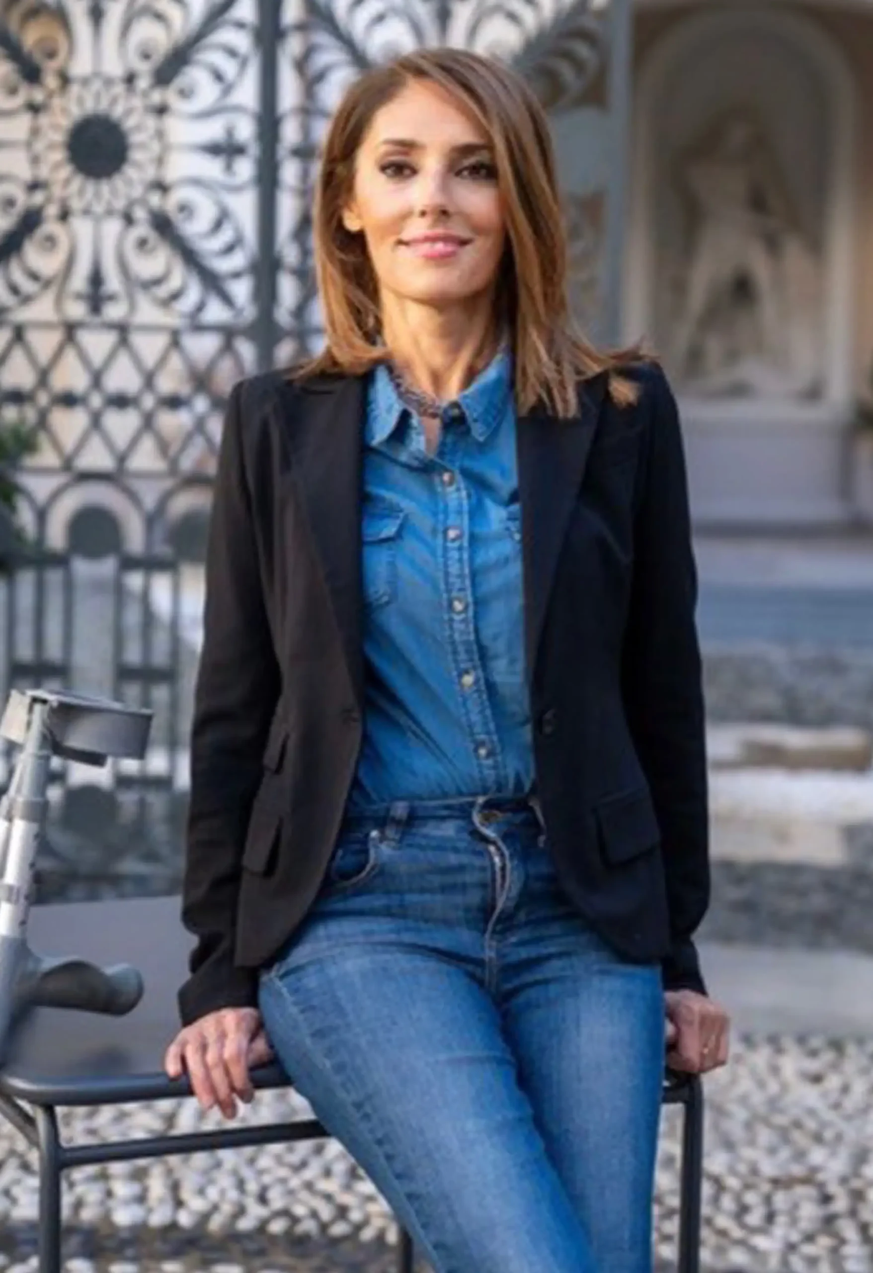 Professional portrait of Francesca Martinengo smiling and leaning on a table with crutches, wearing a black blazer, denim shirt, and blue jeans in front of an ornate wrought-iron gate.