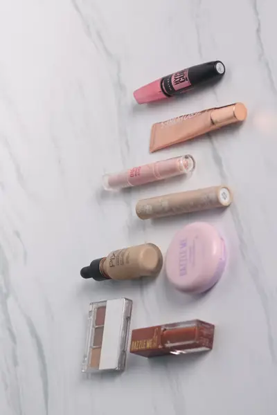 Overhead view of eight makeup products in pink, peach, tan, and brown arranged on a white marble surface, including mascara, a tube, lip and concealer wands, a dropper foundation bottle, a compact, a brow palette, and a lip gloss.