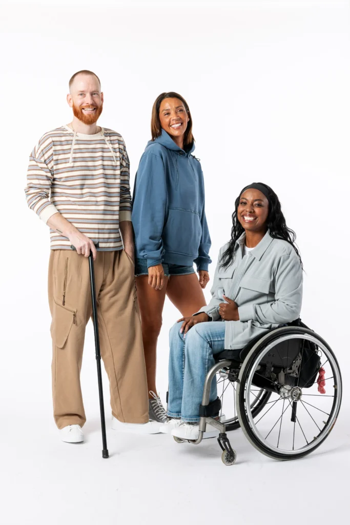 Three smiling individuals standing together on a white background, including a man using a walking cane, a woman in a blue hoodie, and a woman in a wheelchair wearing a grey jacket and jeans.