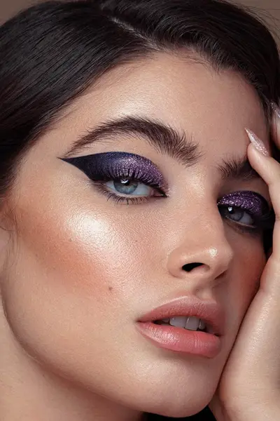Close-up beauty portrait of a woman with dark slicked-back hair, wearing sparkly deep-purple eyeshadow with a dramatic winged liner, a glowing cheek highlight, and glossy peach-pink lips, with one hand touching her face.