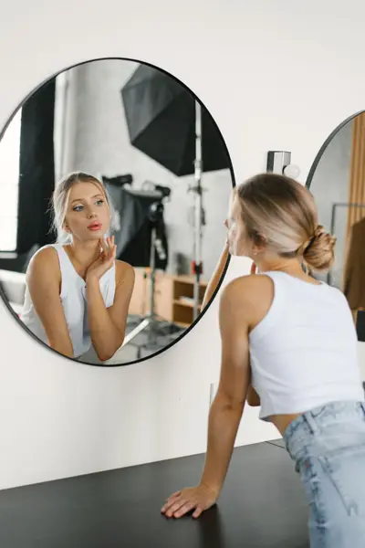 Woman with blonde hair in a low bun, wearing a white tank top and light-wash jeans, leans on a dark counter and looks at her reflection in a round black-framed mirror, with photography studio lights visible in the mirror.