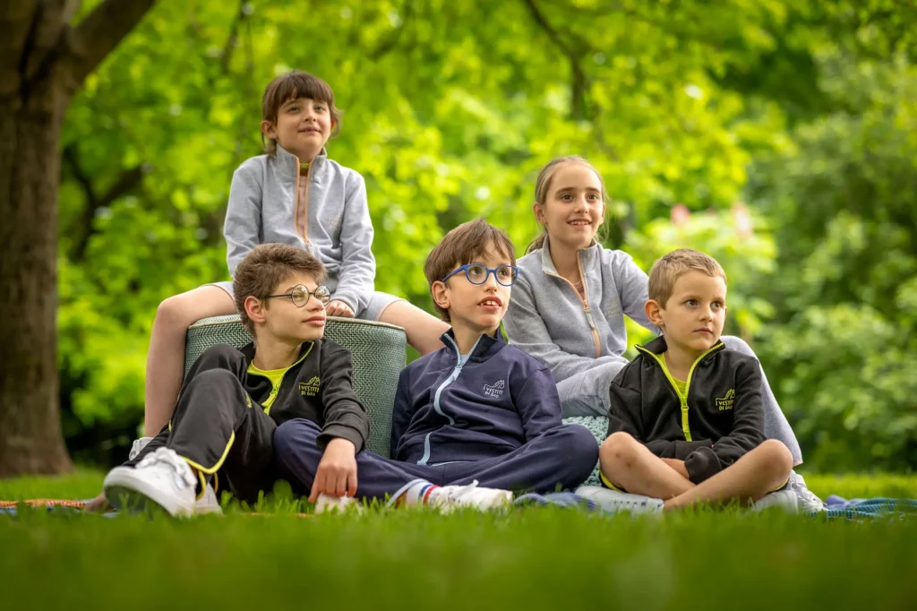 A group of five young children sitting together on a grassy lawn in a park, wearing comfortable gray and navy blue adaptive tracksuits from the I Vestiti di Gio collection.