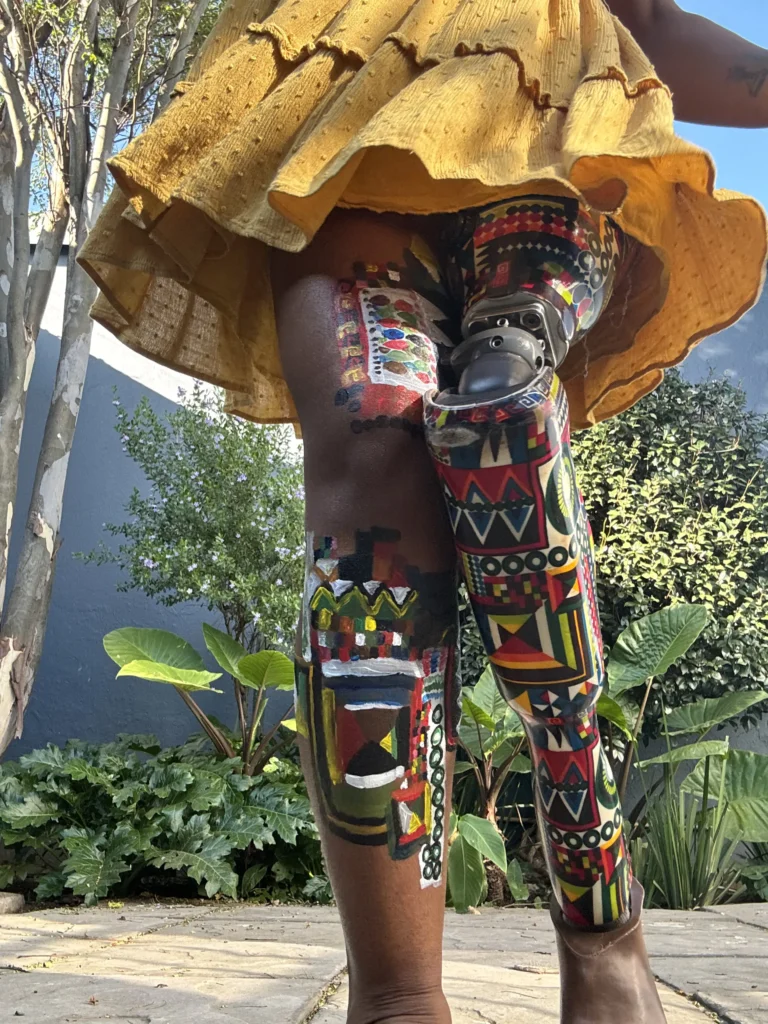 A low-angle close-up shot focusing on a person's legs standing outdoors under a yellow tiered dress. One leg features intricate, colorful body painting with traditional motifs, while the other is a prosthetic leg covered in a matching vibrant, multi-colored geometric African print pattern.