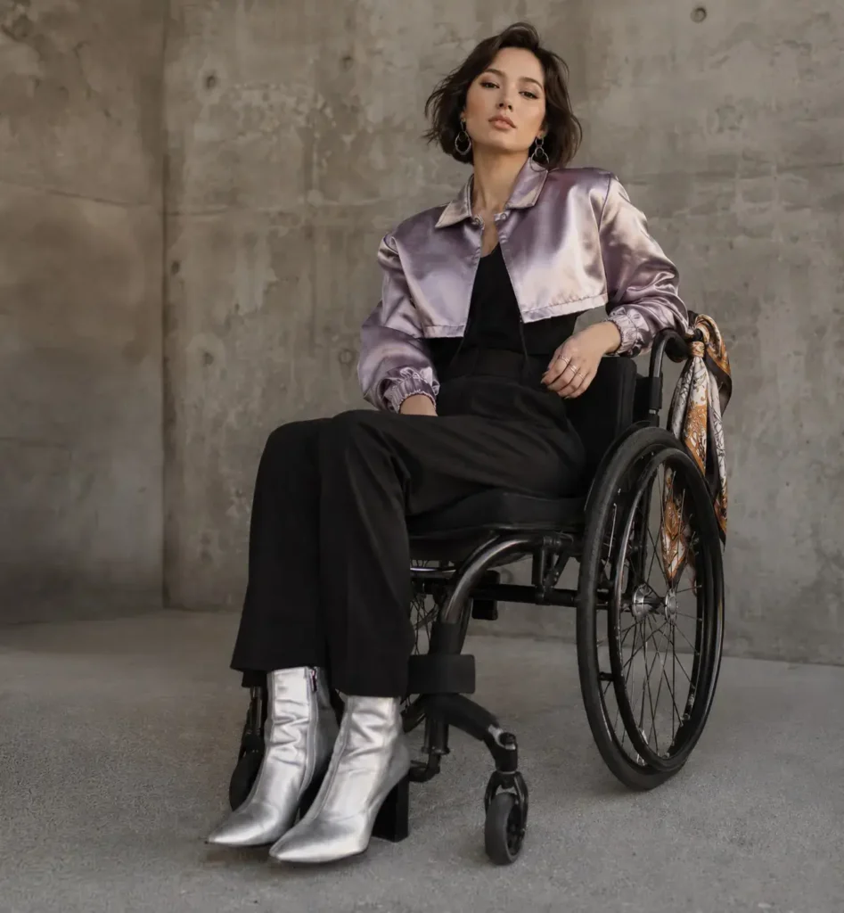 A stylish woman sitting in a wheelchair wearing a cropped metallic lavender satin jacket, black jumpsuit, and pointed silver ankle boots against a concrete wall.