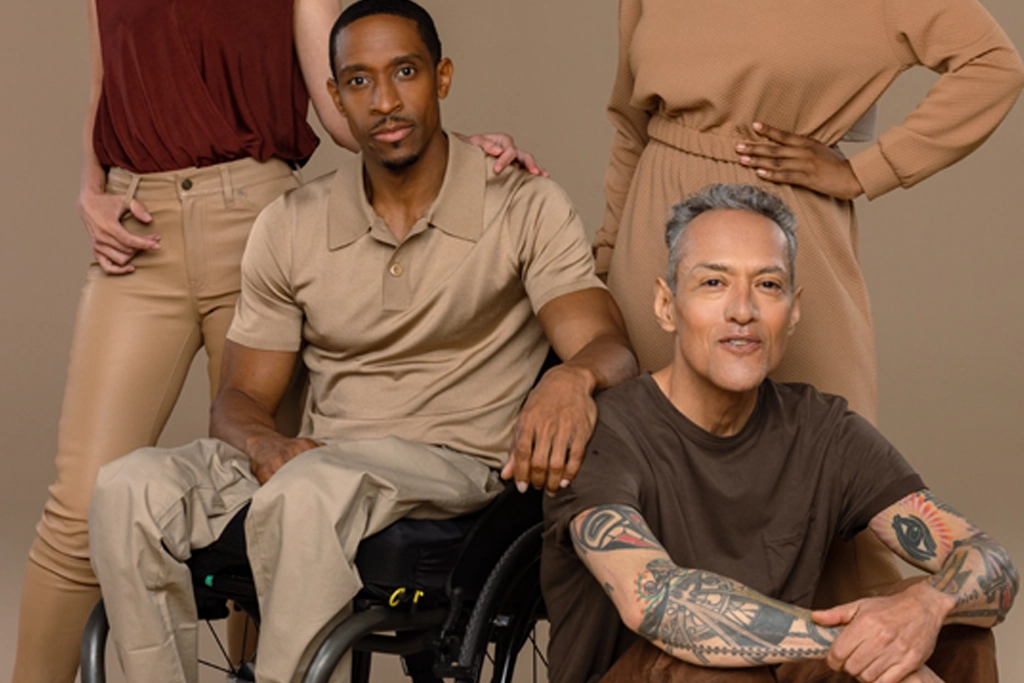 A diverse group of four people posing together in front of a beige background, including a Black man sitting in a wheelchair and a tattooed man sitting on the floor, all dressed in coordinated earth-toned outfits.