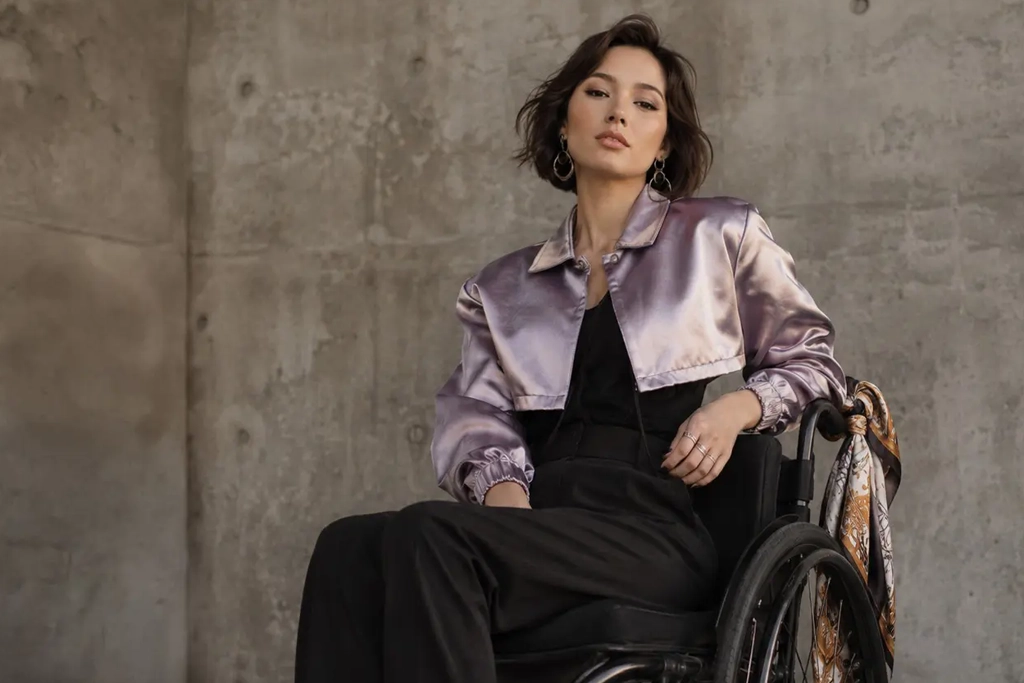A fashionable woman with a short bob haircut sitting in a wheelchair against a concrete wall, wearing a cropped metallic lavender satin jacket over a black top and trousers.