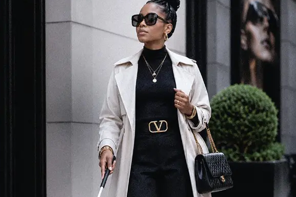 A stylish blind woman walking down a city street while using a white cane, wearing a chic cream trench coat over a black turtleneck, sunglasses, gold jewelry, and carrying a black quilted purse.