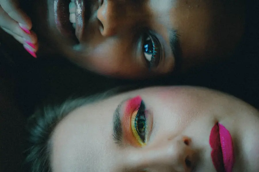 Close-up of two people's faces at close range, one with bright pink lip gloss and pink manicured nails, the other with vivid pink, yellow, and green eyeshadow and bold pink lips.