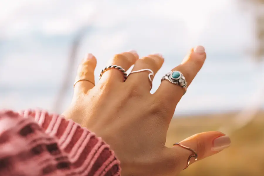 Close-up of a hand wearing multiple silver rings, including one with a turquoise stone, reaching up against a soft blurred outdoor background.