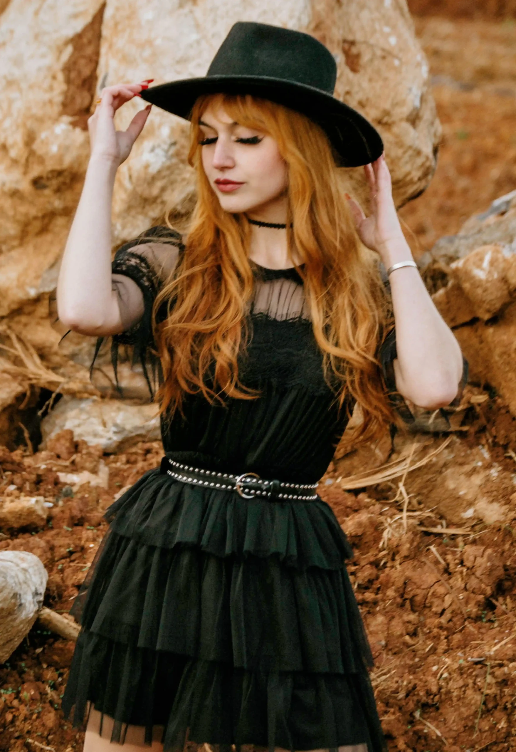 A woman with long red hair wearing a black tulle dress and a wide-brimmed black hat standing in front of a rocky background.