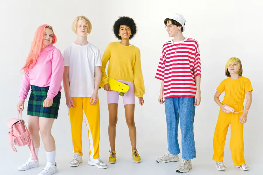 A group of five young people in colorful, trendy outfits pose against a plain white background, one holding a skateboard and another a pink handbag