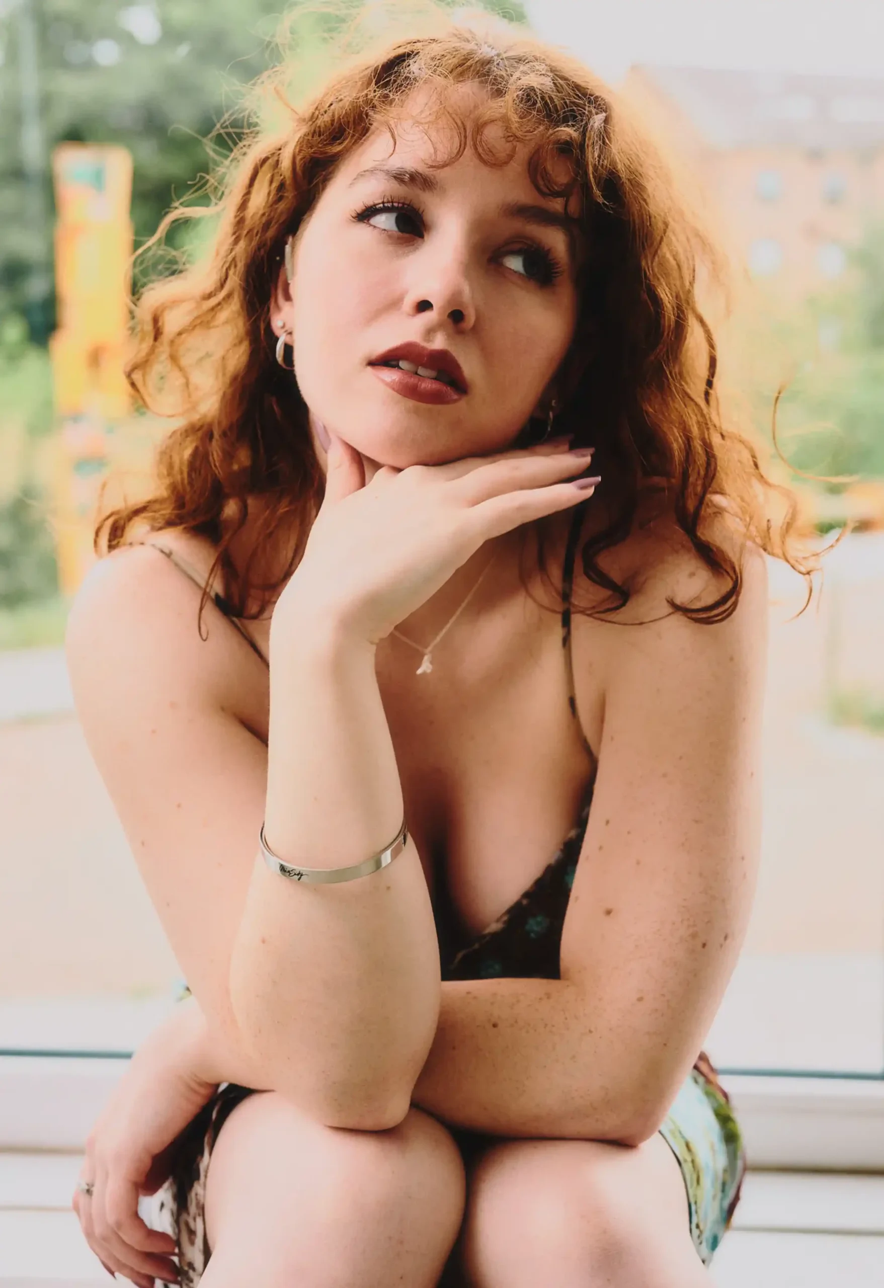 Close-up portrait of a young woman with curly red hair looking off to the side, resting her chin on her hand near a bright window while wearing a delicate silver bracelet.