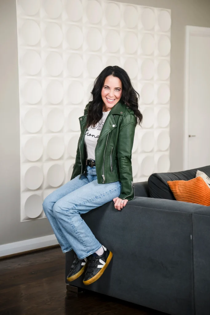 Lainie Ishbia smiling while perched on the edge of a grey sofa, wearing a green leather moto jacket, a white graphic t-shirt, light-wash blue jeans, and black sneakers.
